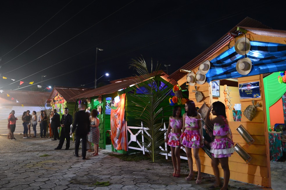 Festejos Juninos no interior com palhoças, bandeirinhas e pessoas dançando em torno da fogueira.