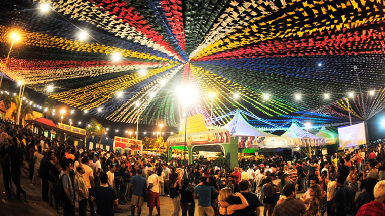 Festas Juninas com pessoas se divertindo em um ambiente típico do interior do Brasil, decorado com bandeirinhas coloridas e barracas de comida.