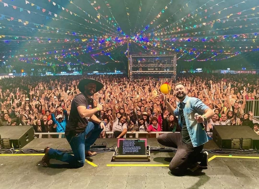 Uma apresentação ao vivo de uma dupla sertaneja em um palco iluminado, rodeados por um público animado.