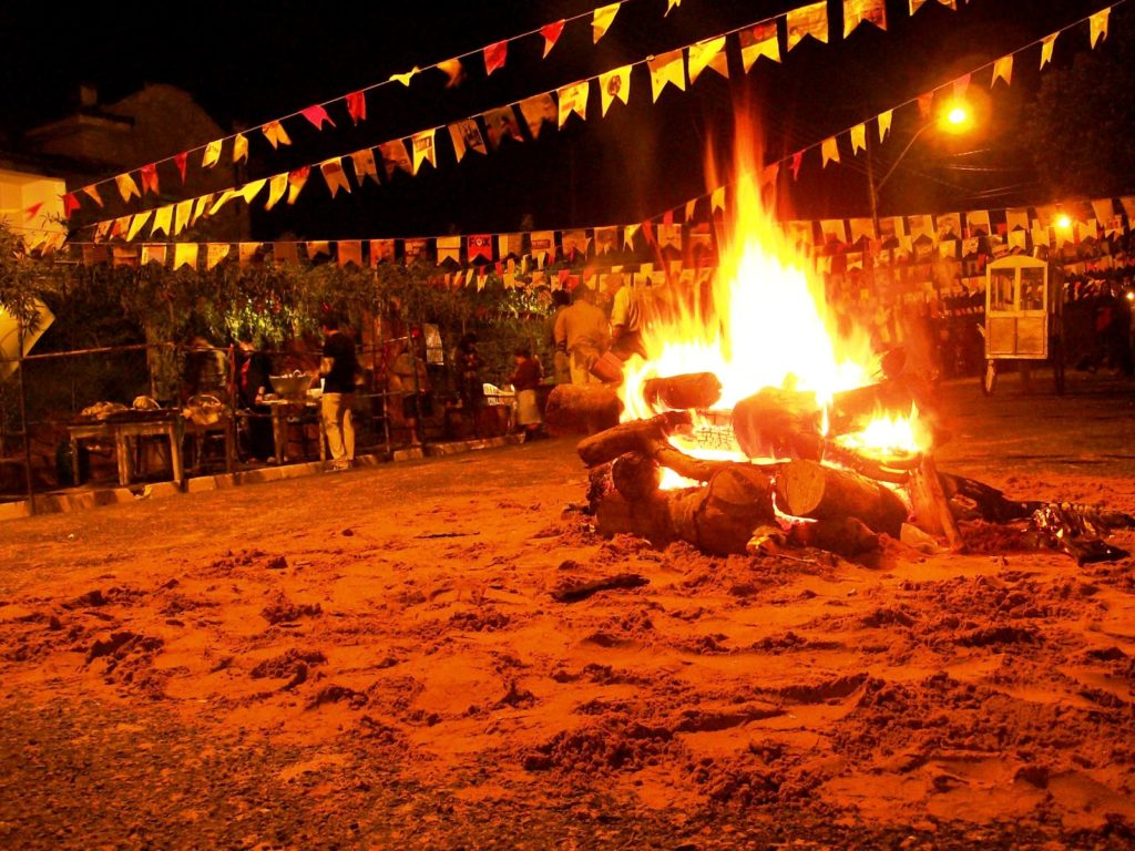 Certa imagem representando uma Festa Junina com bandeirinhas coloridas, fogueira e pessoas dançando quadrilha.