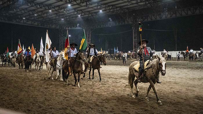 Cavaleiro gaúcho em uma competição de rodeio, cercado por tradições e bandeiras farroupilhas.