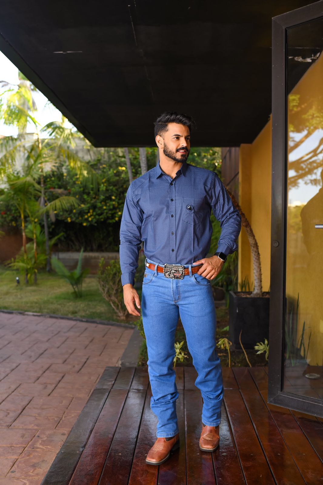 Homem vestindo roupa country, incluindo camisa xadrez, jeans e botas, no ambiente rural.