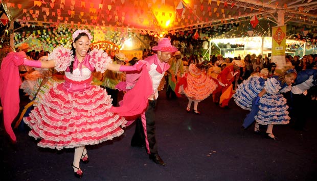 Festa Junina no interior com bandeirinhas coloridas e pessoas dançando ao redor de uma fogueira.