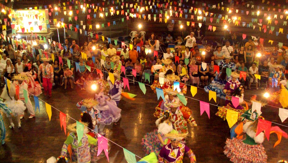 Festa Junina no campo com bandeirinhas coloridas e pessoas dançando quadrilha.