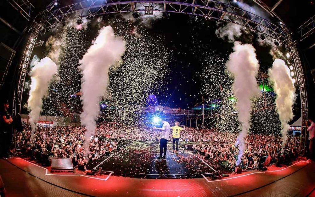 Imagem de um show sertanejo ao ar livre com público animado, bandeirinhas e luzes.